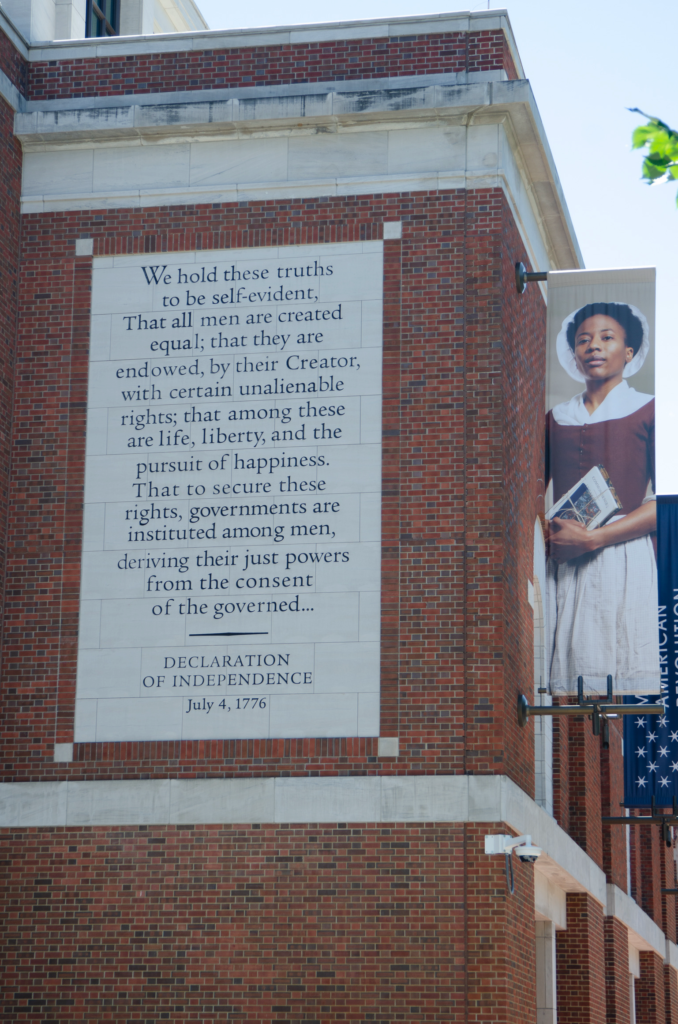 The Declaration of Independence on the side of the Museum of the American Revolution in Philadelphia, next to a banner depicting a black woman (quite likely enslaved) in 18th century clothing.